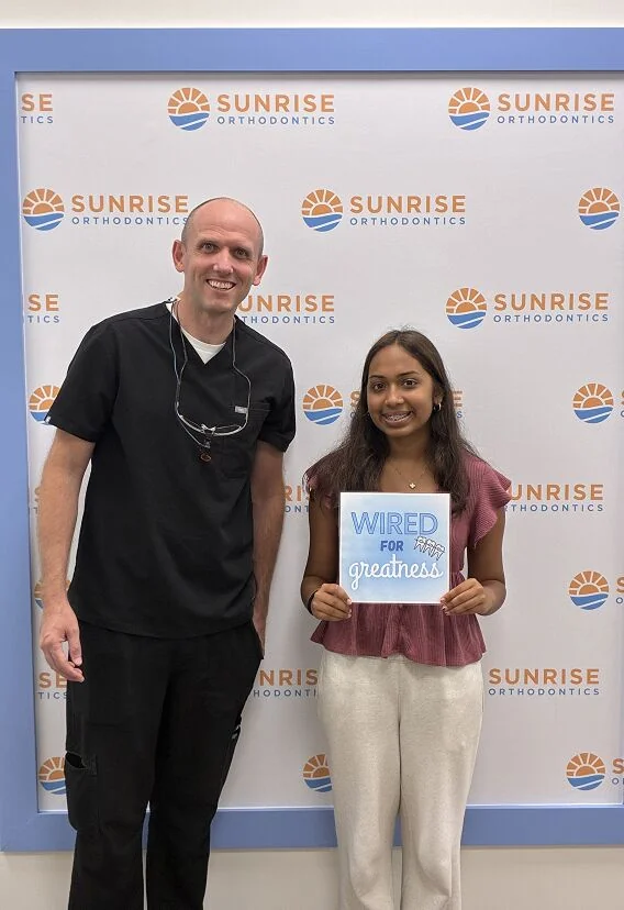 Smiling teen with braces holding a Wired for Greatness sign beside Dr. Steven Dent