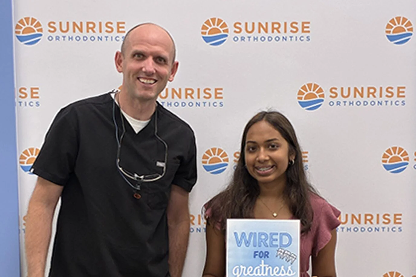 Smiling teen with braces holding a Wired for Greatness sign beside Dr. Steven Dent